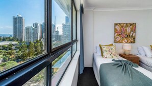 Quarto de hotel com uma janela grande, cama com manta verde, mesa de cabeceira com luminária e um quadro com flores na parede.