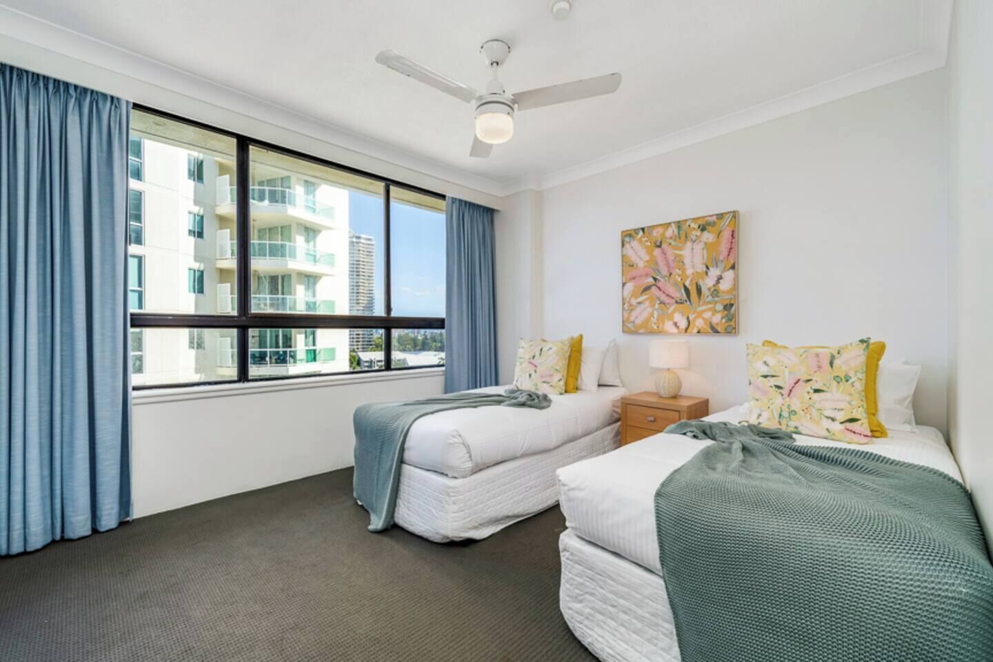 A hotel room with two beds, a ceiling fan, a window with curtains, and a painting on the wall.