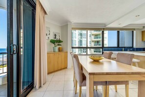 A modern dining area with a large table, chairs, and a view of the sea through large windows. - Carrington Getaway - Ocean Views, Parking, Pool (Main Beach)