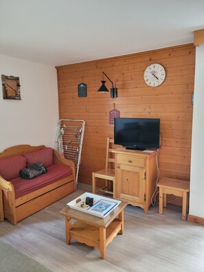 A cozy living room with a wooden sofa, a television, a wooden cabinet, and a wall clock. - QUIET APARTMENT FOR 6 PEOPLE, 45 m², South-Facing Balcony, Pool, Sauna, Steam Room (Valloire)
