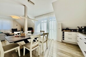 Een moderne keuken met witte kastjes, een eethoek met een houten tafel en stoelen, en een groot raam met dunne gordijnen.