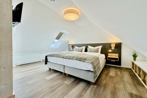 Ein Schlafzimmer mit schrägen Wänden, einem Bett mit Kopfteil, Nachttischlampen, einer kleinen Ablage und einem Fenster.