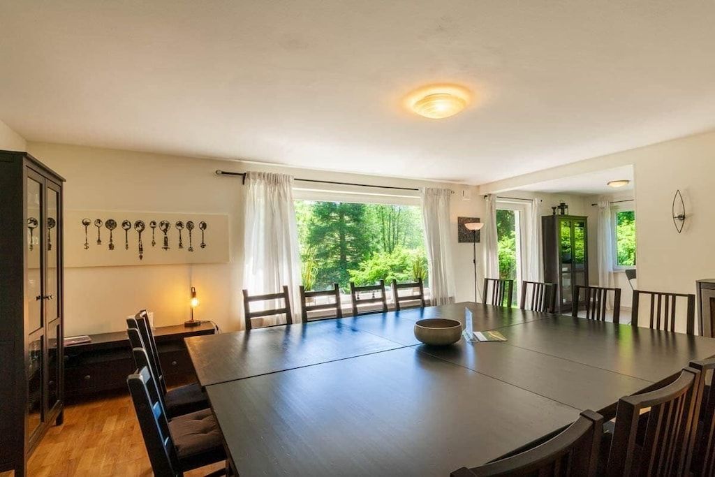 A spacious dining area with a large table and chairs, a view of greenery outside, and a modern light fixture on the ceiling.