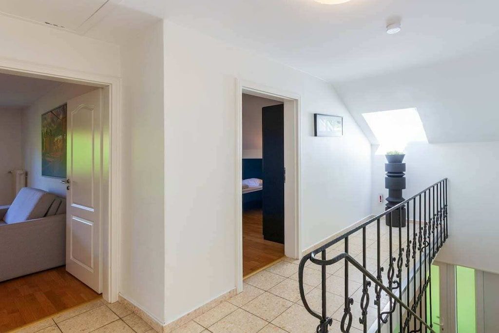 Un hall d’entrée spacieux avec un escalier menant à l’étage supérieur, une porte donnant sur une chambre, et une vue sur une autre chambre avec un lit et une fenêtre.