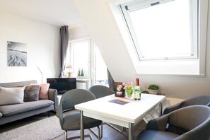 Een moderne woonkamer met een bank, een eettafel en stoelen. Er is een televisie, een raam met gordijnen en een kleine plant op de tafel.