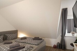 A bedroom with a bed, pillows, a bedside table, a window with curtains, and a plant on the windowsill. - Holiday apartment in Sylt with 2 bedrooms (Wenningstedt)