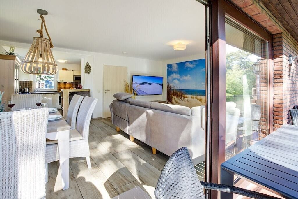Ein modernes Wohnzimmer mit einer Couch, Esstisch und Stühlen, einem Fernseher und einem großen Fenster mit Blick auf den Strand.