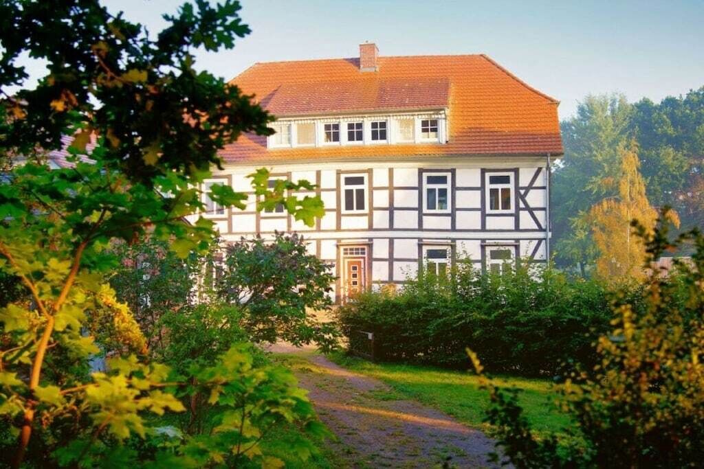 A traditional half-timbered house with a red-tiled roof, surrounded by greenery and a gravel path.