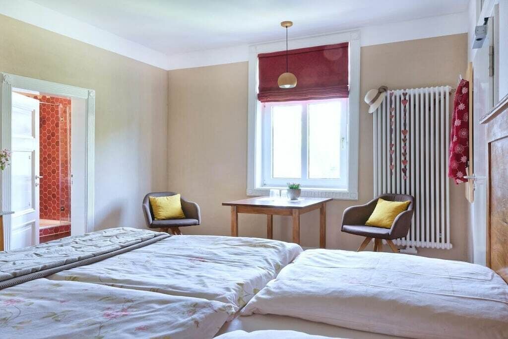 A room with two beds, a small table, two chairs, a window with a red blind, and a radiator.
