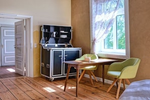 A room with a wooden floor, a dining table with chairs, a kitchenette with cabinets, and a window with curtains. - Holiday room with 1 bedroom (Groß Zecher)