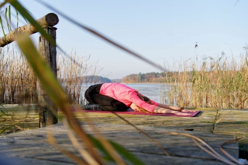 A person performing a yoga pose on a mat by a body of water with reeds in the background.