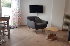 A living room with a grey armchair, a television mounted on the wall, a wooden chair, a small wooden table, and a radiator. - Holiday apartment with 1 bedroom (Dornumersiel)