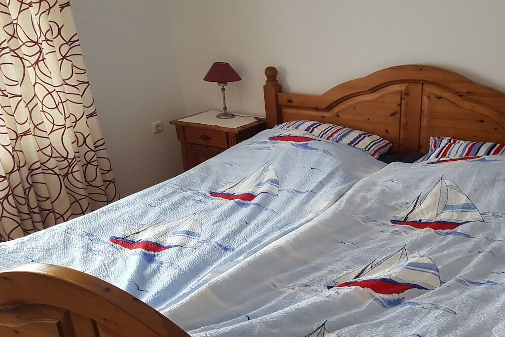 A bed with a sailboat pattern, a wooden headboard, a nightstand with a lamp, and a patterned curtain.