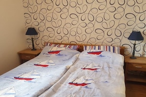 Een bed met een sprei met zeilbootmotief, twee nachtkastjes met lampen, en behangpapier met een patroon.