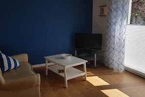 Een woonkamer met een bank, een witte salontafel, een flatscreen televisie en een raam met jaloezieën.