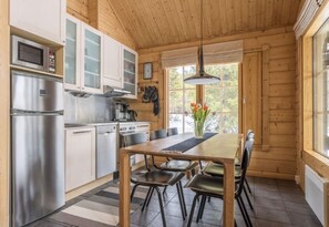 A wooden cabin interior with a kitchen, dining table, chairs, and a window with a view. - Ihtinginrinne | Peaceful 2BR Log Villa by the Lake (Ruka)