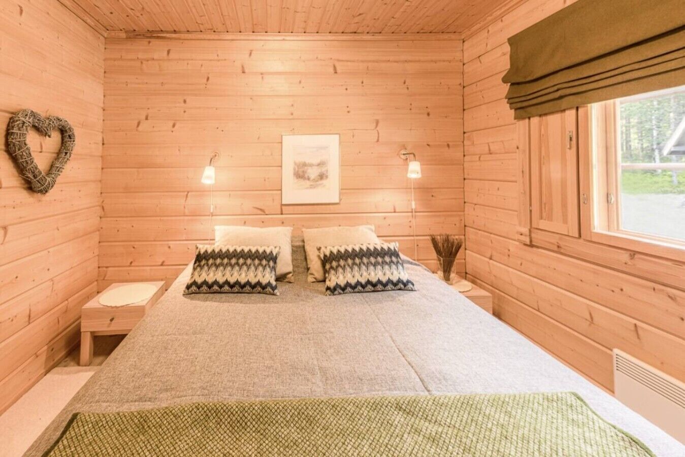 Habitación de cabaña de madera con cama, dos almohadas, un adorno con forma de corazón, un cuadro enmarcado y una ventana.
