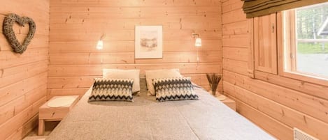 Habitación de cabaña de madera con cama, dos almohadas, un adorno con forma de corazón, un cuadro enmarcado y una ventana.