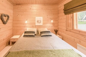 Ein Zimmer einer Holzhütte mit einem Bett, zwei Kissen, einer herzförmigen Dekoration, einem gerahmten Bild und einem Fenster.
