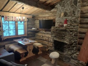 L’intérieur d’un chalet confortable, avec des murs en rondins de bois, une cheminée en pierre, une table à manger avec des chaises et un téléviseur fixé au mur.
