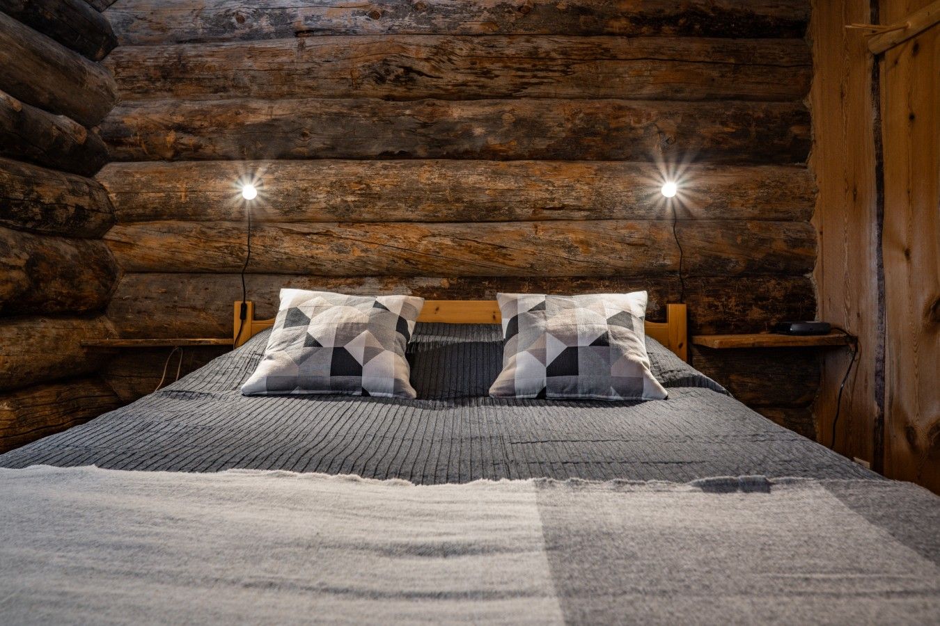 A log cabin bedroom with a bed, two pillows, a wooden headboard, and a nightstand with a lamp.