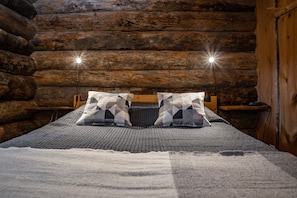 Een slaapkamer in een blokhut met een bed, twee kussens, een houten hoofdbord en een nachtkastje met een lamp.