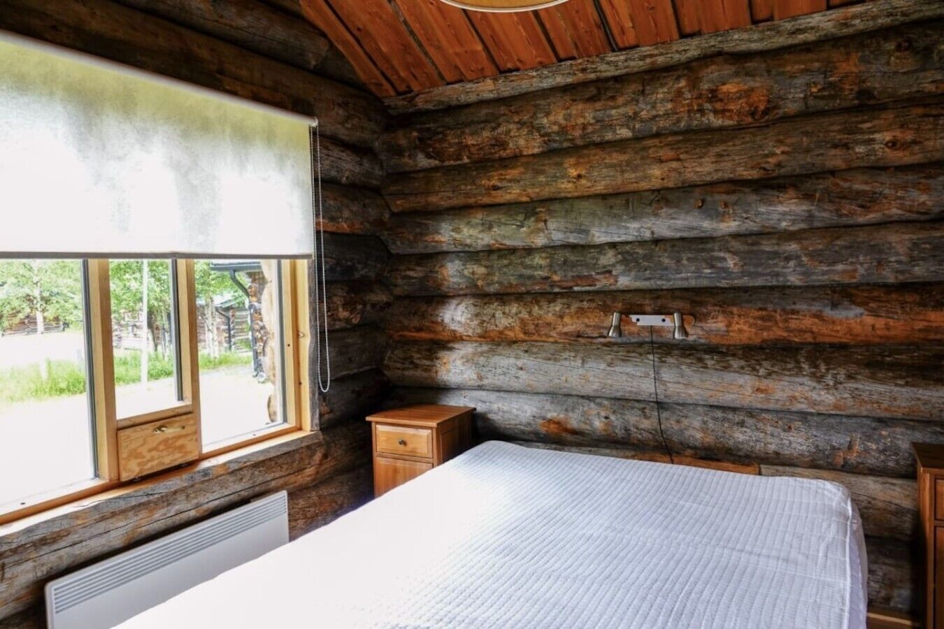 Une chambre dans un chalet en rondins, avec un lit, une petite table de chevet en bois, une fenêtre et un radiateur.