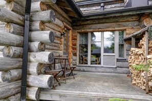 A log cabin with a porch, a stack of firewood, and a view of the surrounding forest.