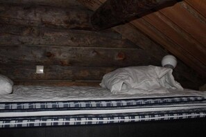 Quarto de sótão pequeno e com pouca luz, com uma cama, teto de madeira inclinado e uma única luminária.