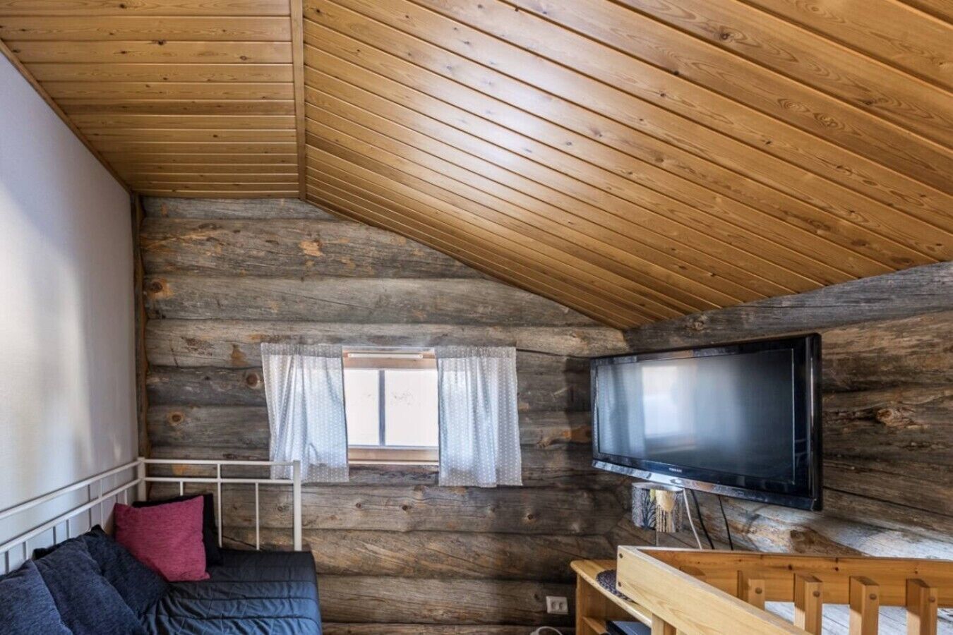 A log cabin interior with a sloped ceiling, a television mounted on the wall, a window with curtains, and a bed with pillows.