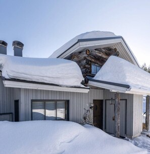A cabin with a sloped roof covered in snow, a garage door, and a wooden log structure. - Rukaköngäs D 17 | Comfy Apt with Fireplace & Sauna (Ruka)