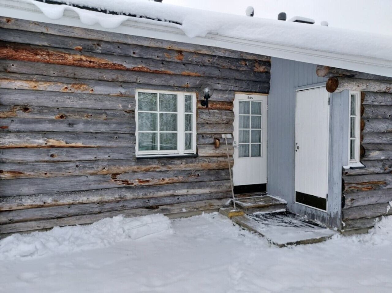 A rustic log cabin with a snow-covered roof, a front door, and a window.
