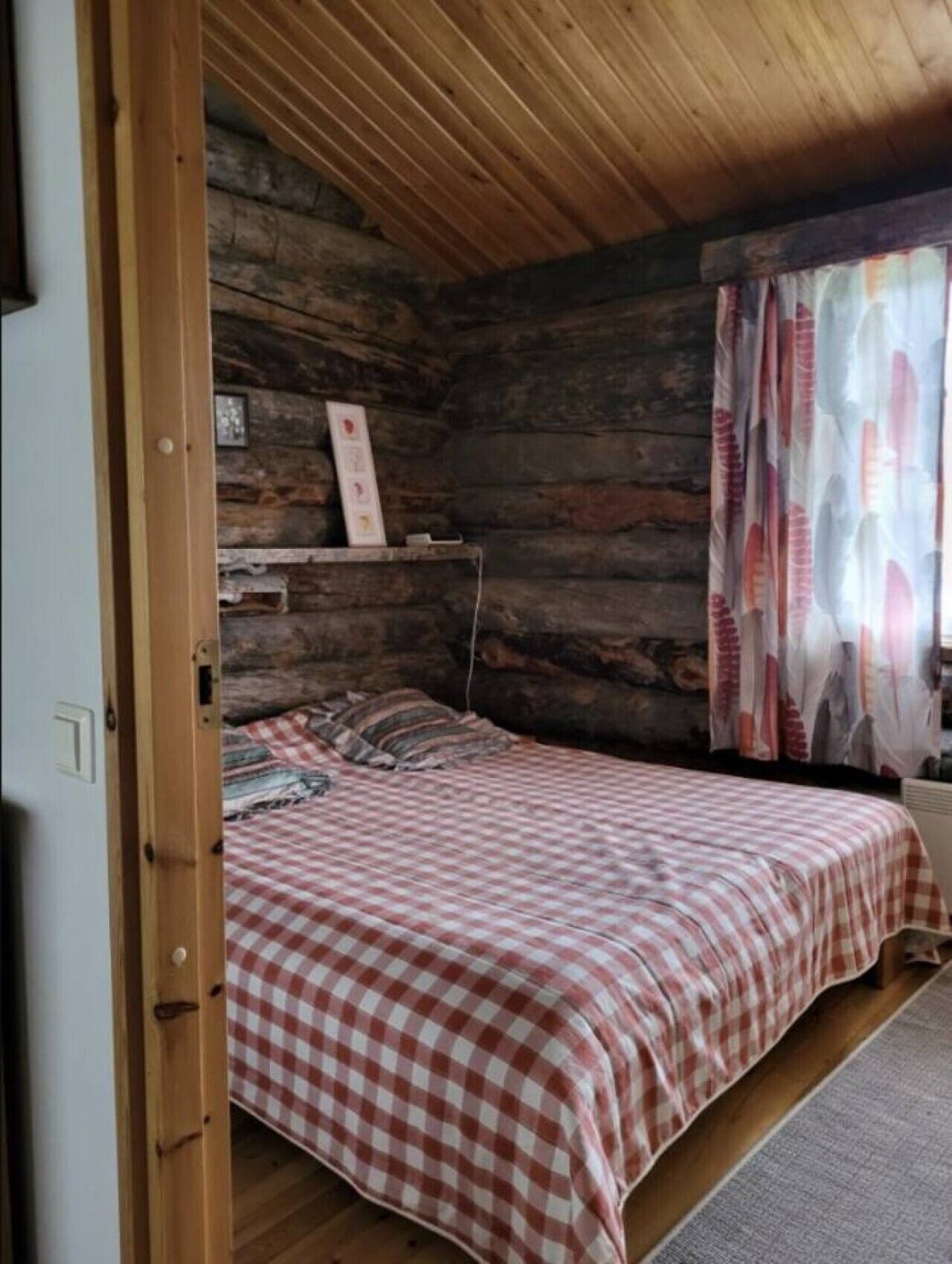 Een slaapkamer met een geruite sprei, houten wanden, een raam met gordijnen en een plank met een ingelijst beeld.