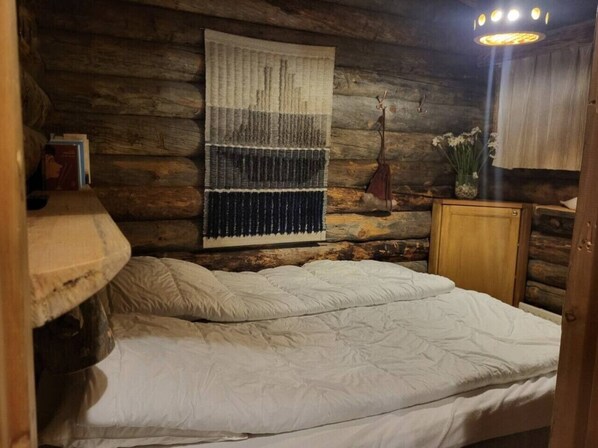 Une pièce rustique en bois, avec un lit, une tapisserie accrochée au mur, une petite armoire et une plante suspendue.