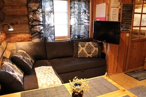 A cozy cabin interior with a sofa, patterned cushions, a flat-screen TV, and a window with a view of trees.