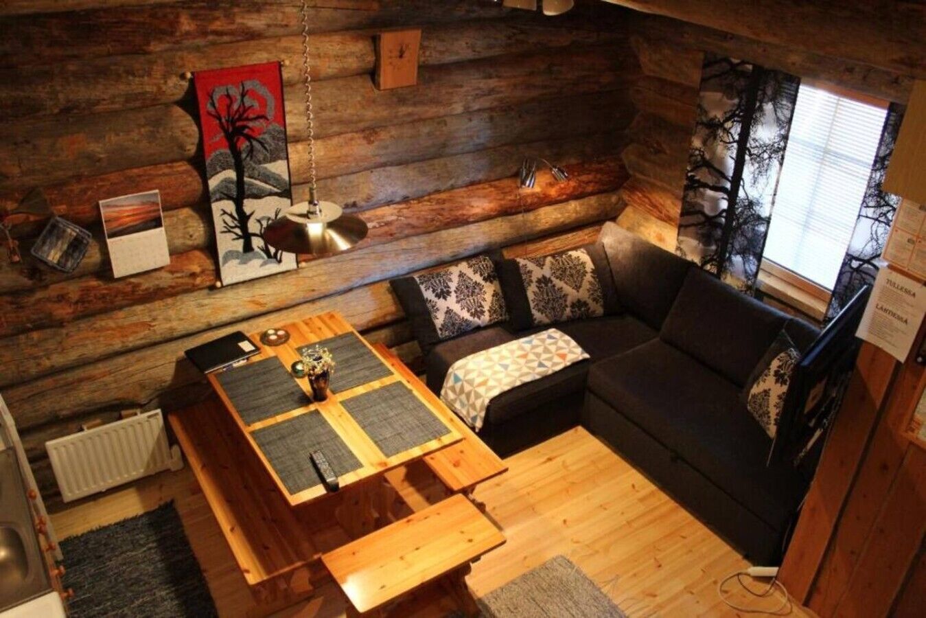 A cozy cabin interior with a wooden dining table, a sofa with patterned cushions, and a hanging light fixture.