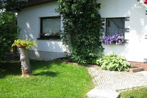 A white building with two windows, one with a flower box containing purple flowers and another with a black frame. There is a tree with green leaves partially obscuring the window, and a brick pathway leading to the building. - Holiday home with 2 bedrooms (Muldenhammer\/OT Hammerbrücke)