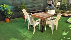 Área externa com mesa de madeira e cadeiras de plástico branco em grama sintética, cercada por plantas em vasos.