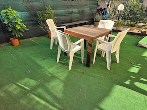 Ein Außenbereich mit Sitzgelegenheiten, einem Holztisch und wei ßen Plastikstühlen auf Kunstgrün, umgeben von Topfpflanzen.