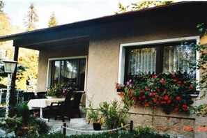 A building with a covered patio area, a table with chairs, and a window with red flowers. - Eismann Holiday Home (Auerbach OT Schnarrtanne)