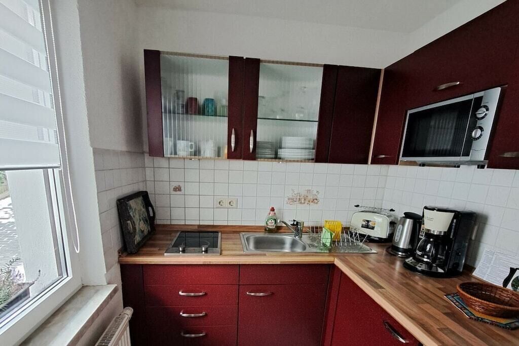 Una cocina equipada con microondas, tostadora y cafetera. Se ve una ventana con persianas, un lavamanos y armarios.