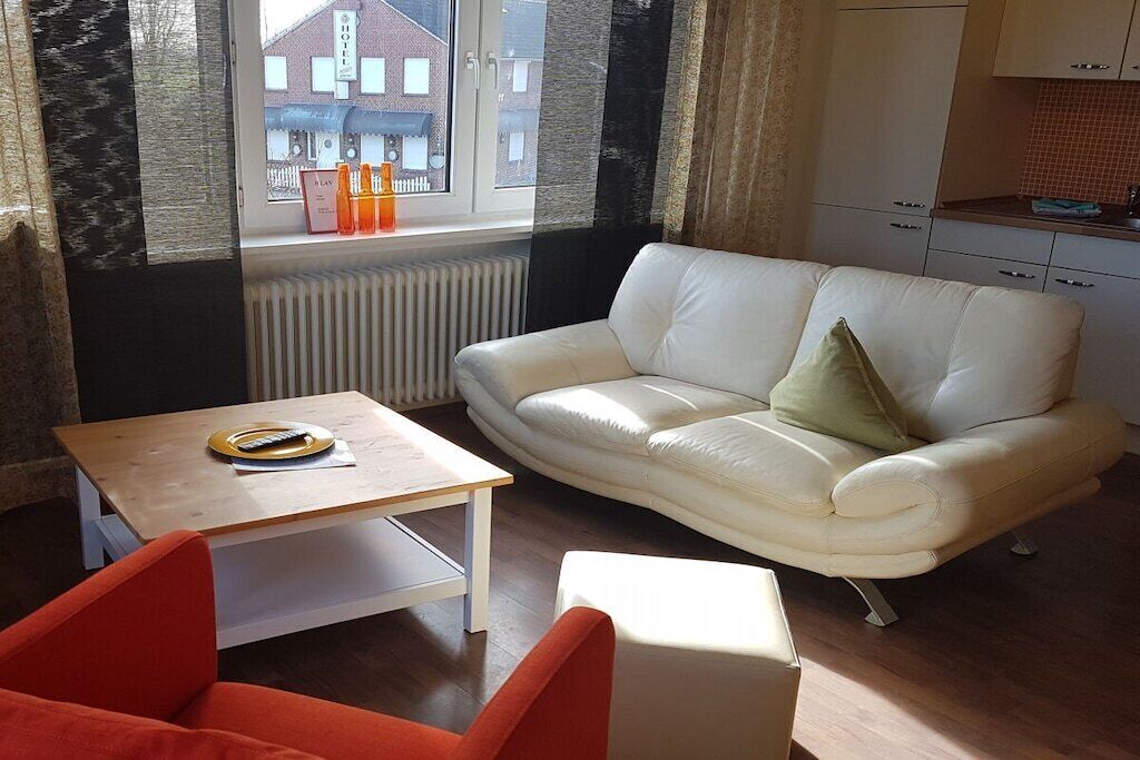 A living room with a white sofa, a wooden coffee table, and a red armchair. A window with a view of buildings is visible.