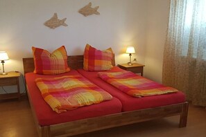 A double bed with a red bedspread and two matching pillows, flanked by nightstands with lamps, in a room with a simple decor and a curtain on the window. - Holiday apartment with 1 bedroom (Dornumersiel)