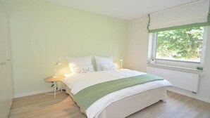 Quarto com cama, mesa de cabeceira, abajur e janela com vista para o verde.