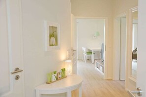 Un couloir avec une porte blanche, une petite table ornée de quelques objets décoratifs, un tableau encadré au mur et, au fond, un coin repas.