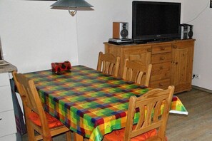 Een eetruimte met een tafel, bedekt met een kleurrijke tafelkleed, houten stoelen, een televisie op een houten kast en een lamp.