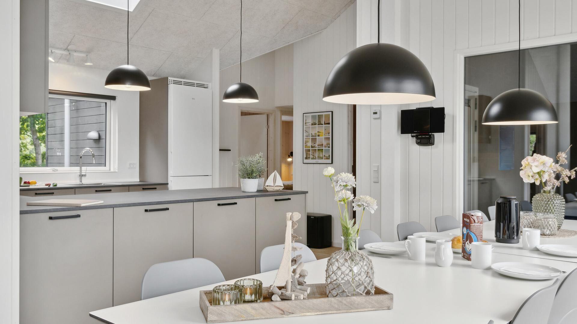 Cozinha moderna com iluminação pendente, área de jantar com mesa branca posta para dois e uma janela ampla que oferece luz natural.