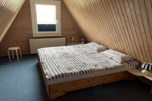 A bedroom with a sloped ceiling, a window, a bed with striped bedding, a bedside table with a lamp, and a small stool. - Holiday home with 2 bedrooms (Spieka-Neufeld (Wurster Nordseeküste))