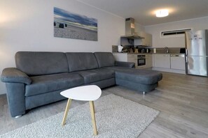 A modern living room with a grey sectional sofa, a white coffee table, and a kitchen area with white cabinets and a stainless steel refrigerator. - Holiday apartment with 1 bedroom (Dierhagen)