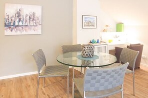 A dining area with a round glass table and four chairs, a cityscape painting on the wall, and a kitchen area in the background. - Holiday apartment with 2 bedrooms (Timmendorfer Strand)